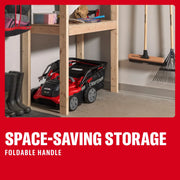 Photo of Craftsman lawn mower CMCOE210P2 with folded handle stored under wooden shelf in garage, next to boots and tools.