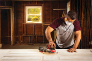 Photo of person using Craftsman CMCW220B cordless sander on a wooden door in a workshop.